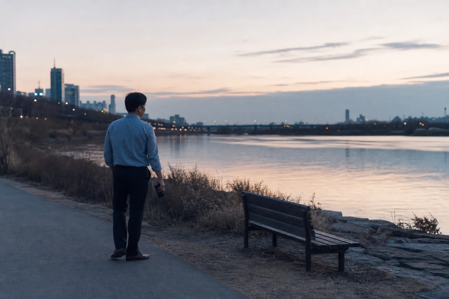 A tired worker pausing beside a river at dusk with a phone lowered at their side